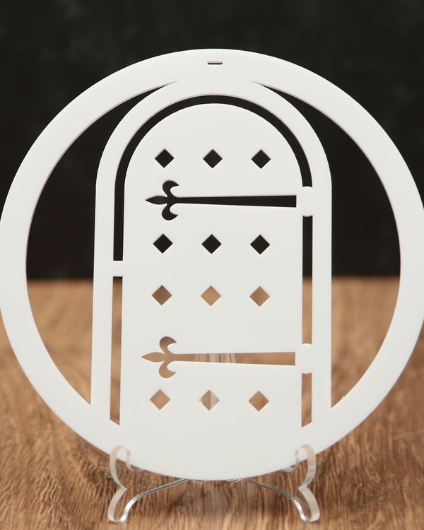 A white laser-cut acrylic Christmas ornament shaped like a circular frame with an arched door design, displayed on a clear acrylic stand on a wooden surface against a dark background.