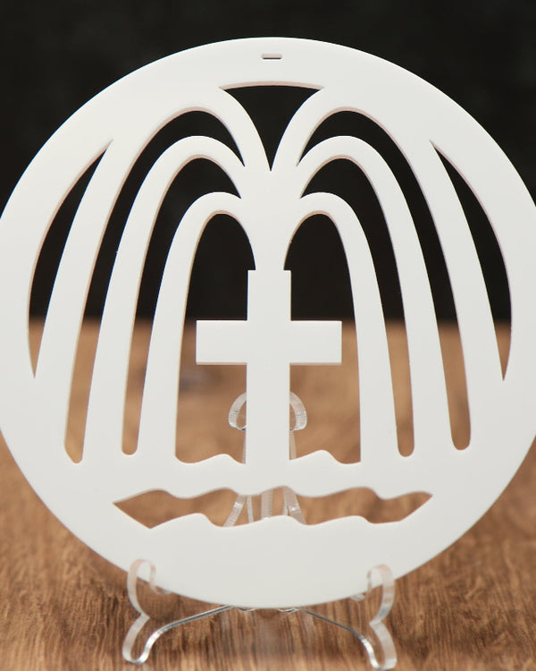 A white circular Christmas ornament with a cross in the center and fountains gushing from the cross, displayed on a clear stand on a wooden surface against a dark background.