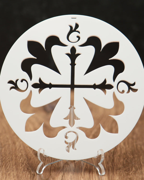 A round white Christmas ornament with an ornamental cross and floral cutout design, displayed on a small clear stand on a wooden surface against a dark background.