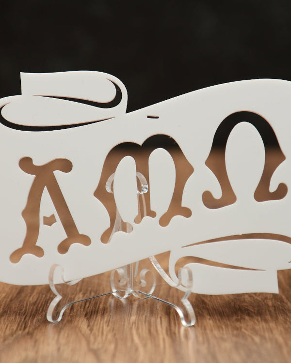 A white laser-cut acrylic scroll sign featuring the Greek letters Alpha, Mu and Omega, displayed on a clear acrylic stand over a wooden surface with a dark background.