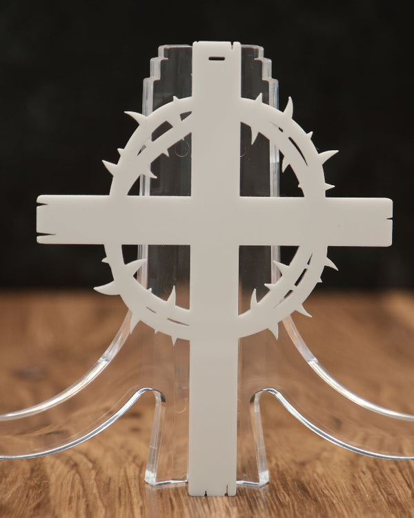 A white laser-cut decorative cross with a crown of thorns motif and a clear acrylic stand, displayed on a wooden surface against a dark background.