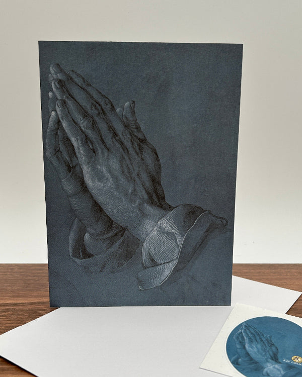 A dark slate greeting card of praying hands displayed on a wooden table with a white envelope and a small circular sticker.