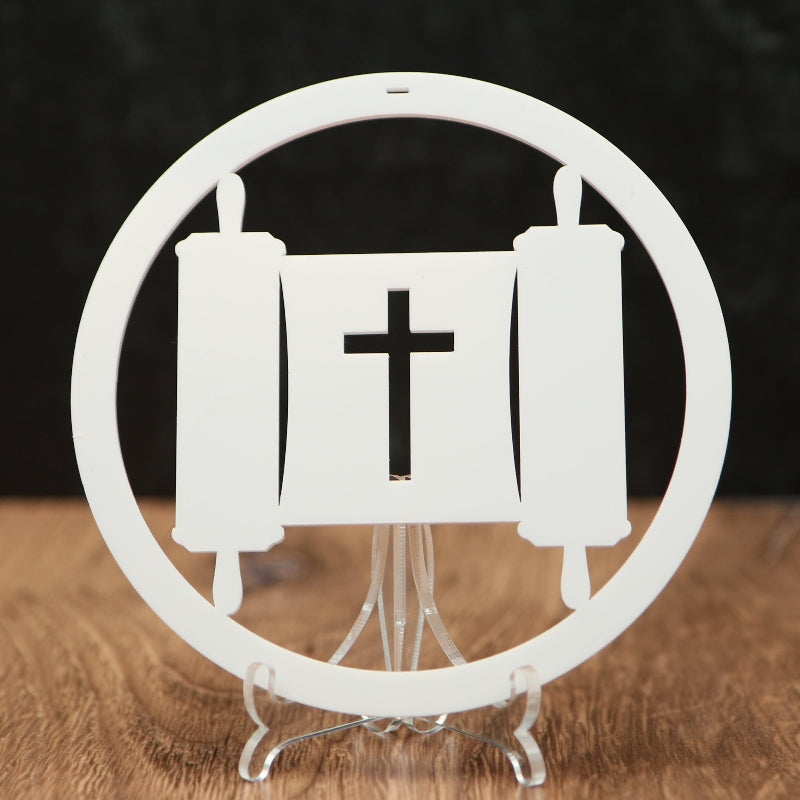 A white circular decorative Christmas ornament with a cross in the center and scroll motifs, displayed on a clear stand over a wooden surface against a dark background.