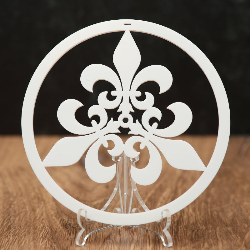 A white circular Christmas ornament with a fleur-de-lis design, displayed on a clear stand over a wooden surface against a dark background.