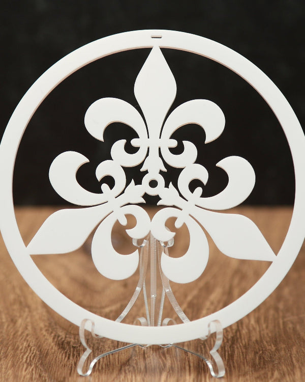 A white circular Christmas ornament with a fleur-de-lis design, displayed on a clear stand over a wooden surface against a dark background.