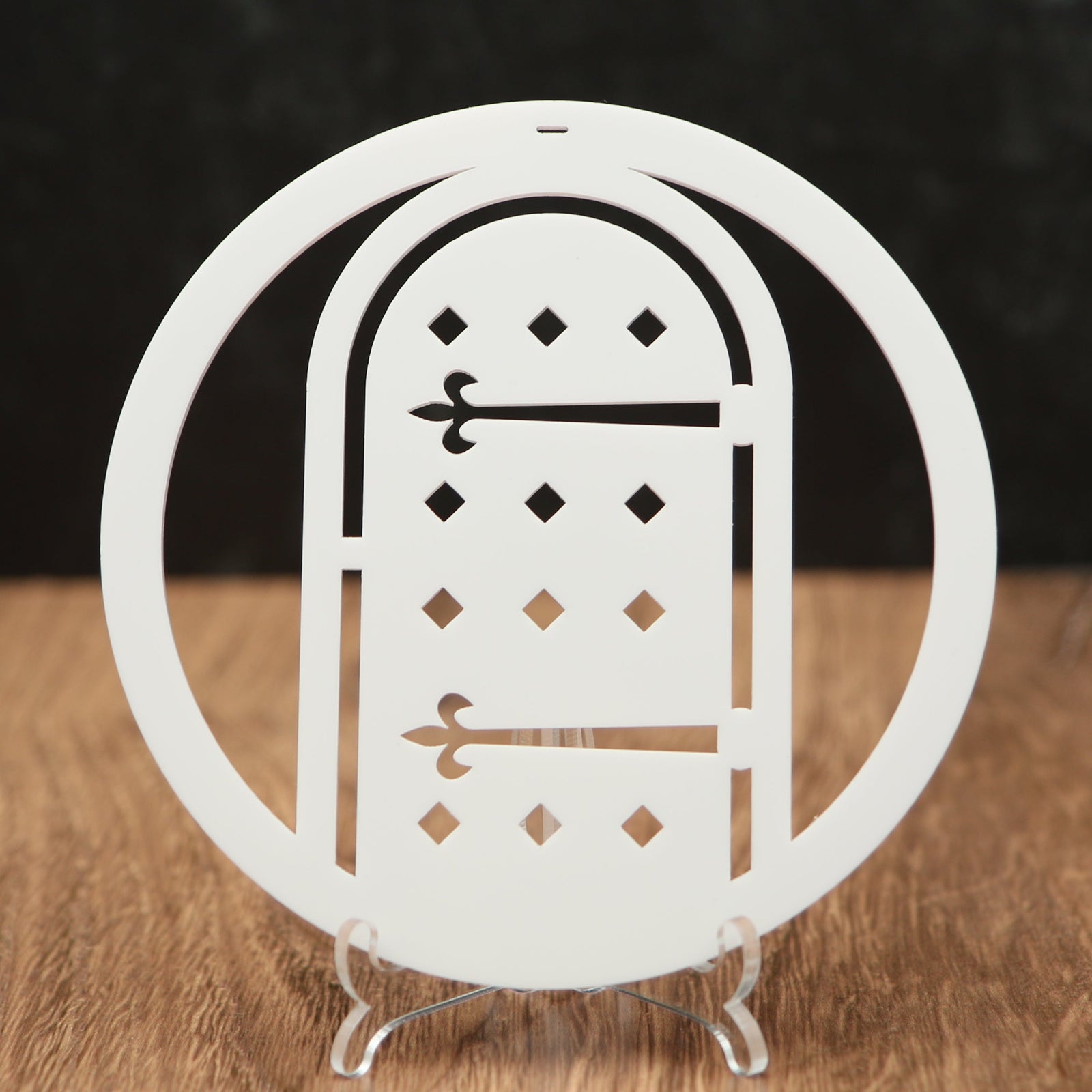A white laser-cut acrylic Christmas ornament shaped like a circular frame with an arched door design, displayed on a clear acrylic stand on a wooden surface against a dark background.