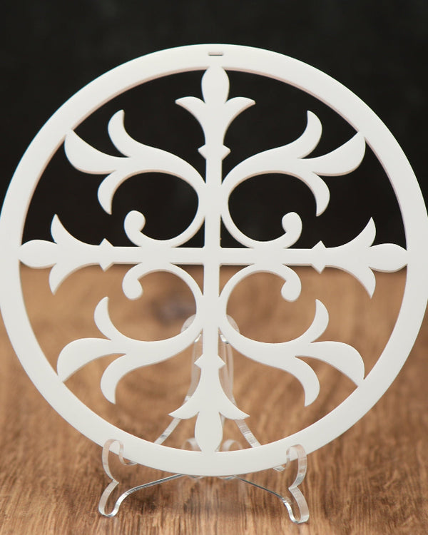 A white circular Christmas ornament with an ornate cutout cross, displayed on a clear stand over a wooden surface against a dark background.