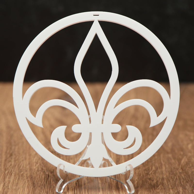 A white Christmas ornament with  a central fleur-de-lis motif, displayed on a small clear stand over a wooden surface against a dark background.
