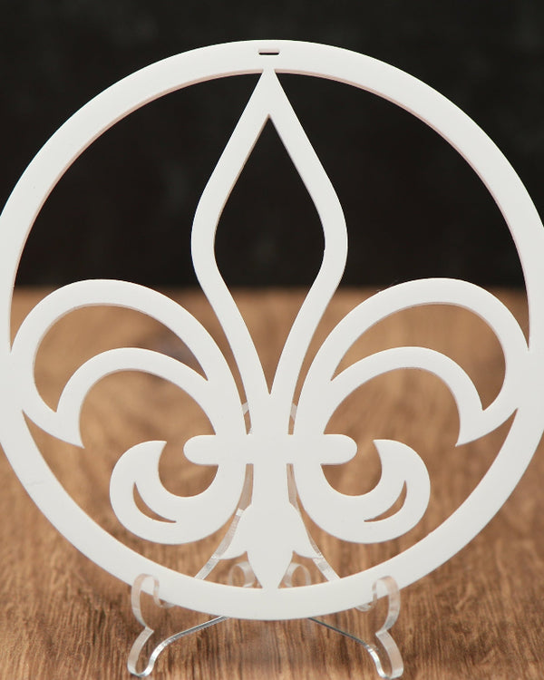 A white Christmas ornament with  a central fleur-de-lis motif, displayed on a small clear stand over a wooden surface against a dark background.