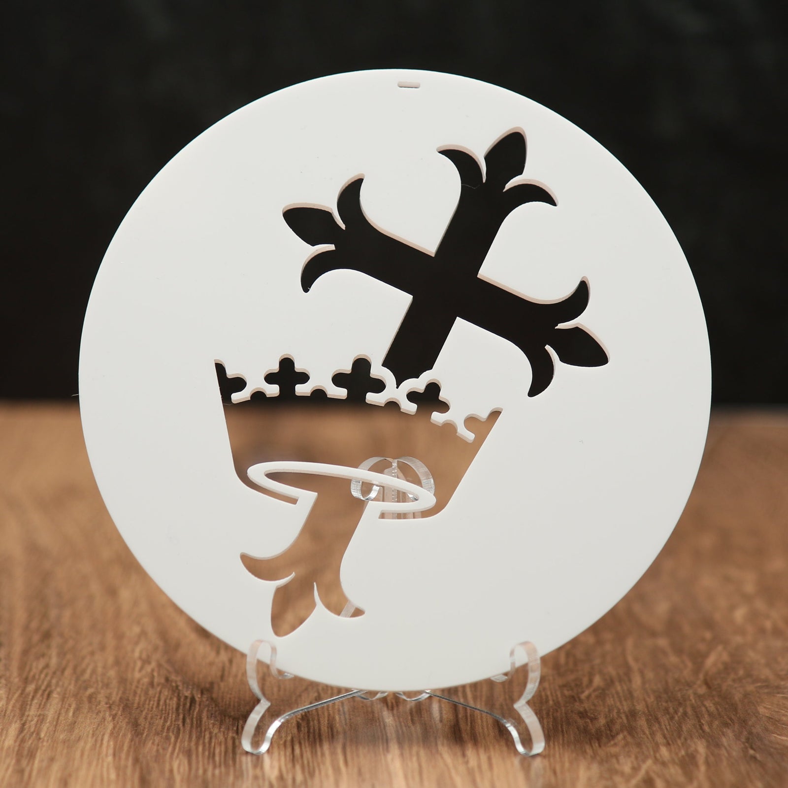 A round white Christmas ornament  with a cross and crown, displayed on a clear stand over a wooden surface against a dark background.