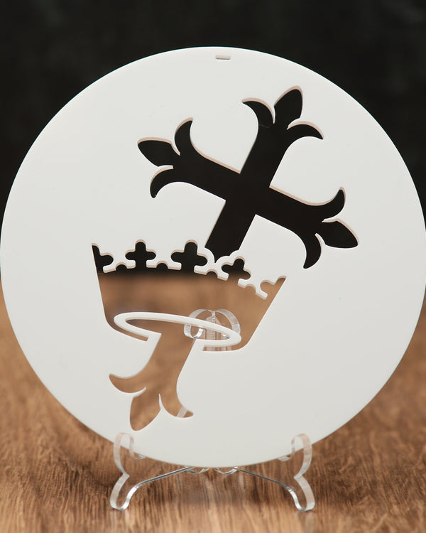 A round white Christmas ornament  with a cross and crown, displayed on a clear stand over a wooden surface against a dark background.