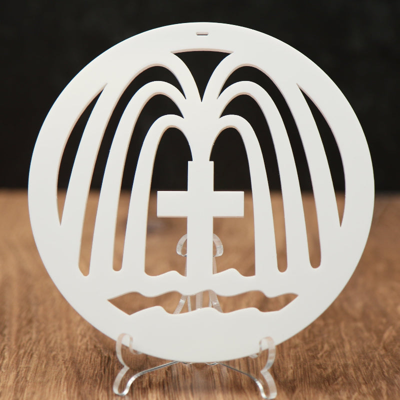 A white circular Christmas ornament with a cross in the center and fountains gushing from the cross, displayed on a clear stand on a wooden surface against a dark background.