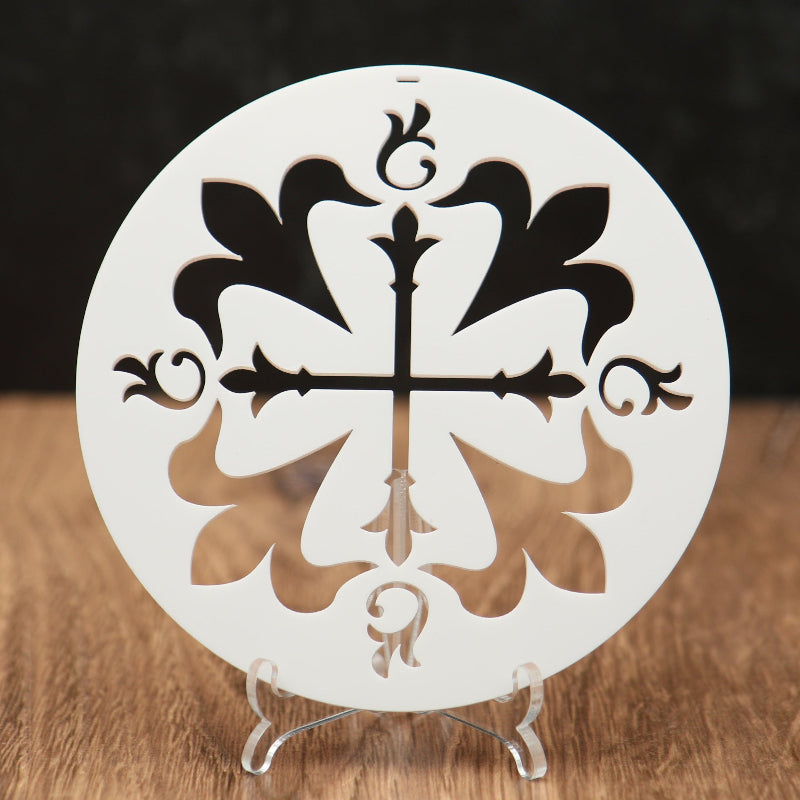 A round white Christmas ornament with an ornamental cross and floral cutout design, displayed on a small clear stand on a wooden surface against a dark background.
