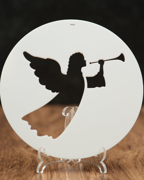 A round white ornament featuring a silhouette of an angel playing a trumpet, displayed on a clear stand on a wooden surface against a dark background.