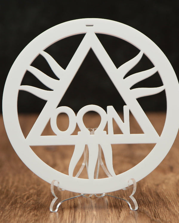A white circular laser-cut ornament featuring a triangle with rays and the word 'OON' in the center, displayed on a clear acrylic stand on a wooden surface against a dark background.
