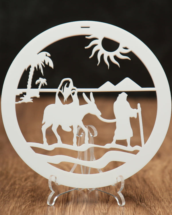 A circular white laser-cut Christmas ornament featuring a desert scene with a camel, figures, mountains, palm tree, and sun, displayed on a clear acrylic stand over a wooden surface against a dark background.