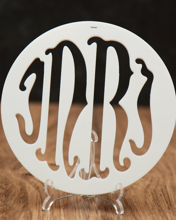 A round white monogrammed acrylic ornament with the initials 'INRI' cut out, displayed on a clear acrylic stand on a wooden surface against a dark background.