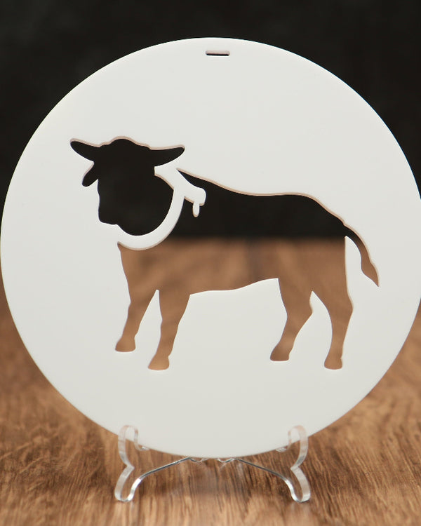 A round white Christmas ornament featuring a bull silhouette cutout, displayed on a clear acrylic stand over a wooden surface against a dark background.