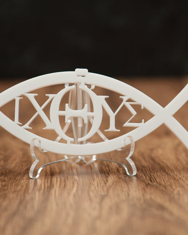 A white fish-shaped Christmas ornament with the Greek letters 'IXOYΣ' cut out in the center, displayed on a clear stand over a wooden surface with a dark background.