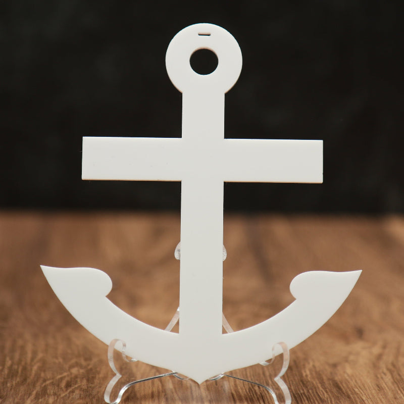 A white acrylic anchor-shaped Christmas ornament displayed on a clear stand on a wooden surface with a dark blurred background.