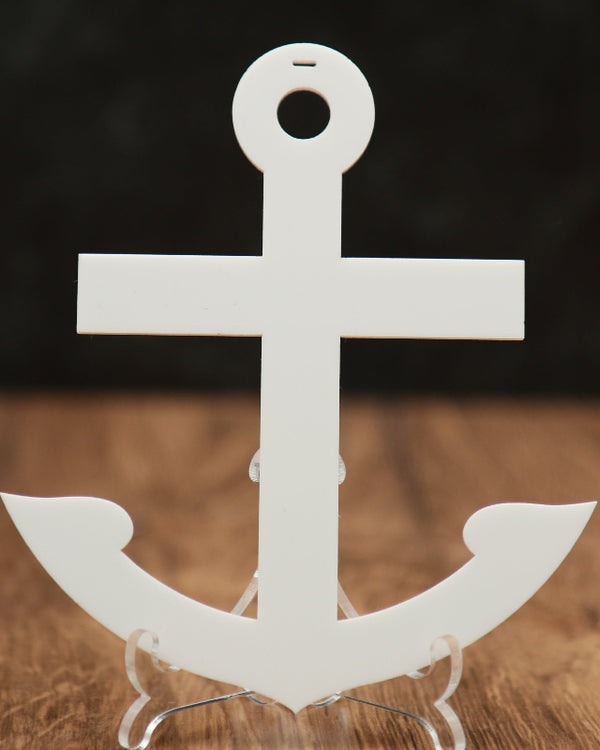A white acrylic anchor-shaped Christmas ornament displayed on a clear stand on a wooden surface with a dark blurred background.