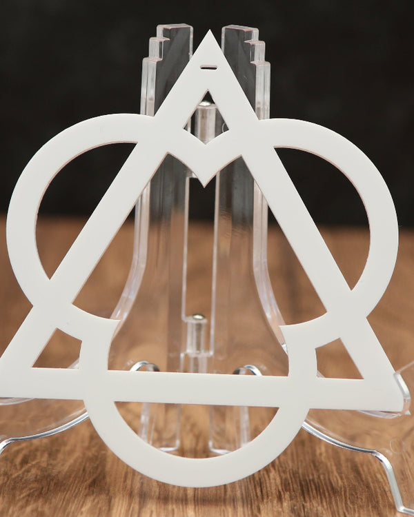 A clear acrylic Christmas ornament featuring a white triangular and circular geometric cutout design, set on a wooden surface against a dark background.