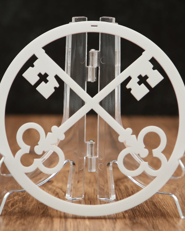 A white circular acrylic stand featuring two crossed decorative keys, displayed on a clear acrylic easel over a wooden surface against a dark background.