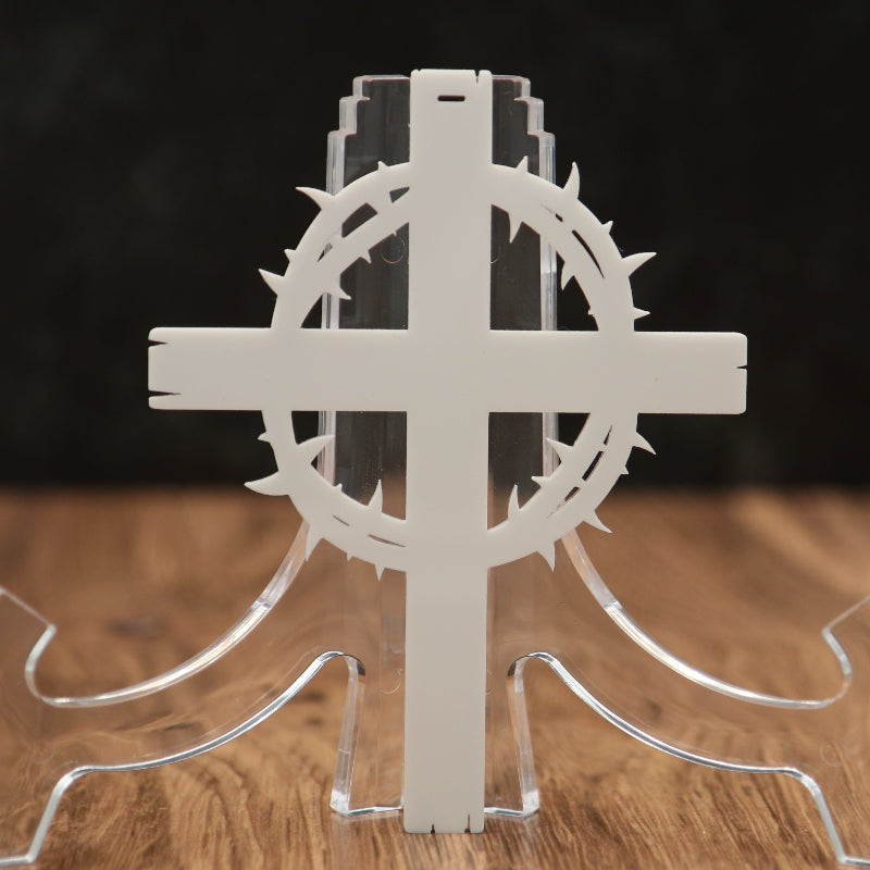 A white laser-cut decorative cross with a crown of thorns motif and a clear acrylic stand, displayed on a wooden surface against a dark background.