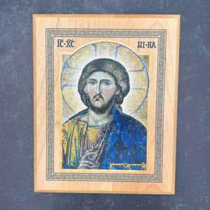 A rectangular wooden icon plaque featuring a painted portrait of Jesus Christ with a gold halo and blue robe, set against a dark textured background.