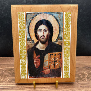 A framed religious icon of Jesus Christ Pantocrator on a wooden easel stand. The icon features a painted portrait of Jesus with a gold halo and a book of Gospels, bordered by ornate gold and white scrollwork on a light oak wood frame, set against a dark textured background.