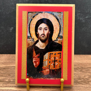 A small framed religious icon of Jesus Christ standing on a gold-colored easel stand, placed on a wooden surface against a dark textured wall.