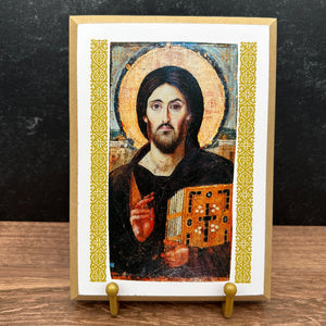 A framed religious icon of Jesus Christ Pantocrator on a wooden tabletop against a dark textured wall. The icon features a gold halo, dark robes, and an ornate gold patterned border, displayed on a small gold stand.