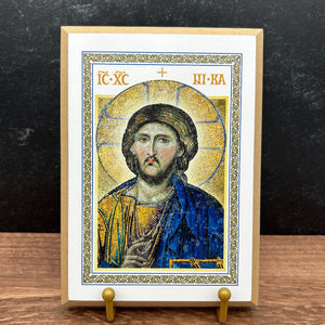 A framed Byzantine-style religious icon of Jesus Christ on a small wooden display stand, photographed against a dark textured wall and wood surface.
