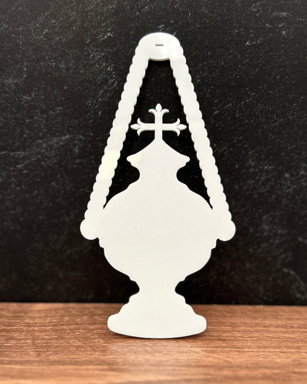 A white decorative silhouette ornament shaped like a classic oil lamp with a cross, standing on a wooden surface against a dark textured background.