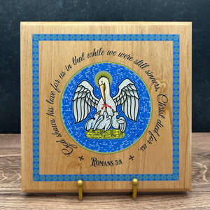 A square wooden plaque with a colorful printed design featuring a pelican, a blue mosaic-style border, and the text 'God's words his love for us in that while we were still sinner Romans 5:8' displayed on a stand against a dark textured background.