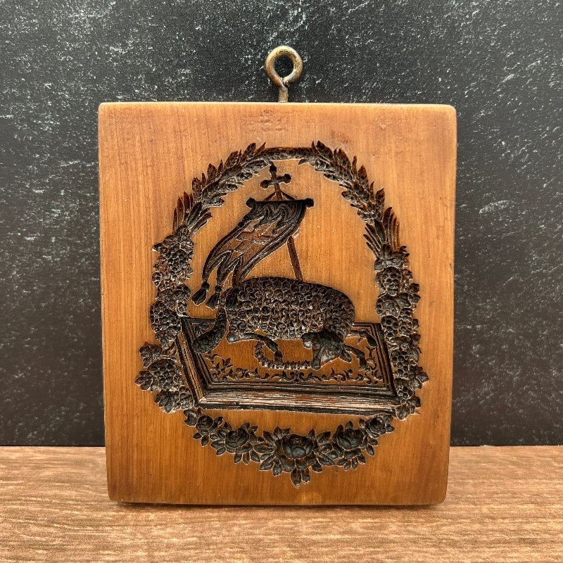 A square wooden box with a carved relief design of a peacock within a floral wreath, featuring a small metal ring on the left side, set against a dark textured background.