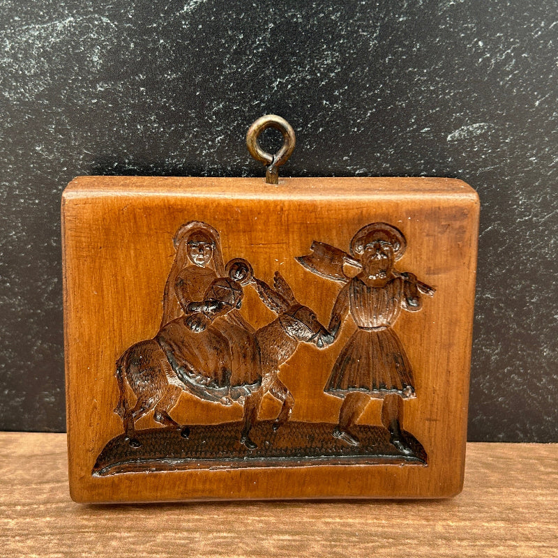 A small rectangular carved wooden plaque or box with a hanging loop, featuring embossed figures, photographed on a dark textured surface next to a wood-grain panel.