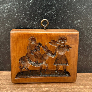 A small rectangular carved wooden plaque or box with a hanging loop, featuring embossed figures, photographed on a dark textured surface next to a wood-grain panel.