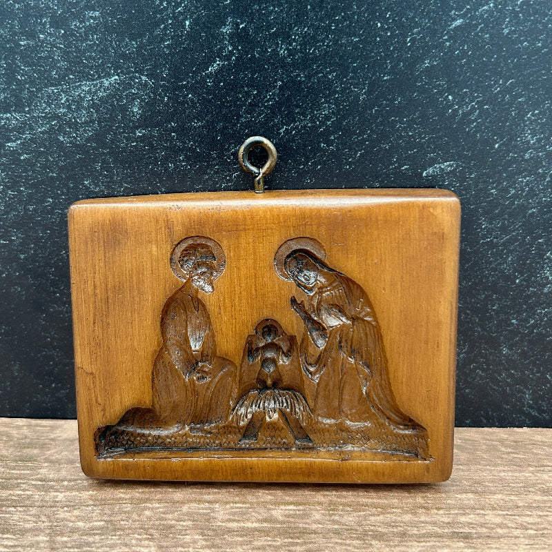 A rectangular wooden nativity cookie mold featuring a carved relief of the Holy Family, displayed on a wooden surface against a dark textured background.