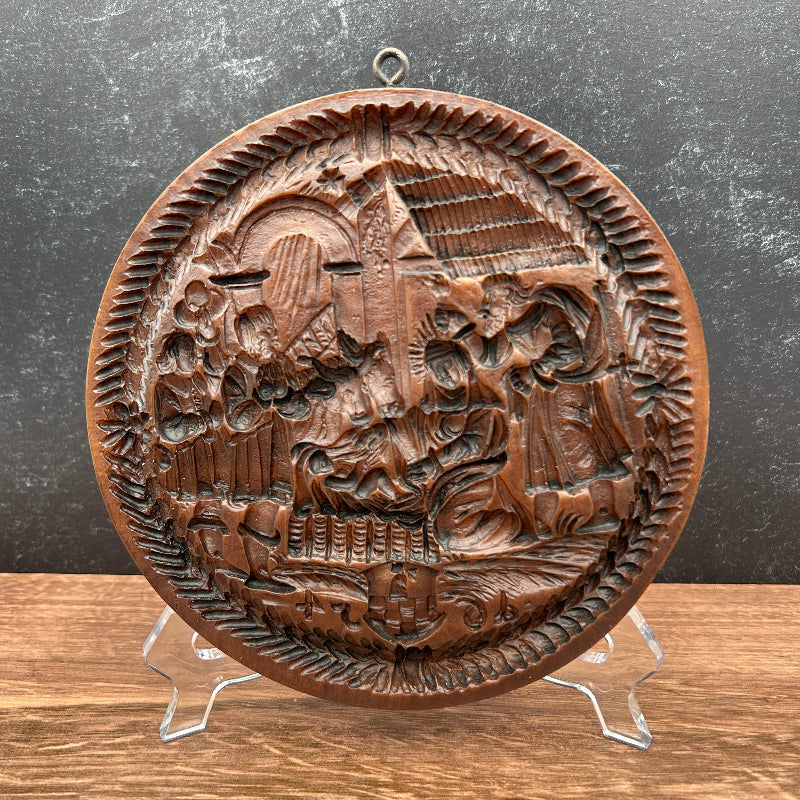 A round, hand-carved cookie mold featuring a detailed historical battle scene, displayed on a clear plastic stand against a split dark gray and wood-grain background.