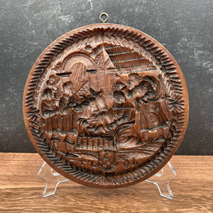 A round, hand-carved cookie mold featuring a detailed historical battle scene, displayed on a clear plastic stand against a split dark gray and wood-grain background.