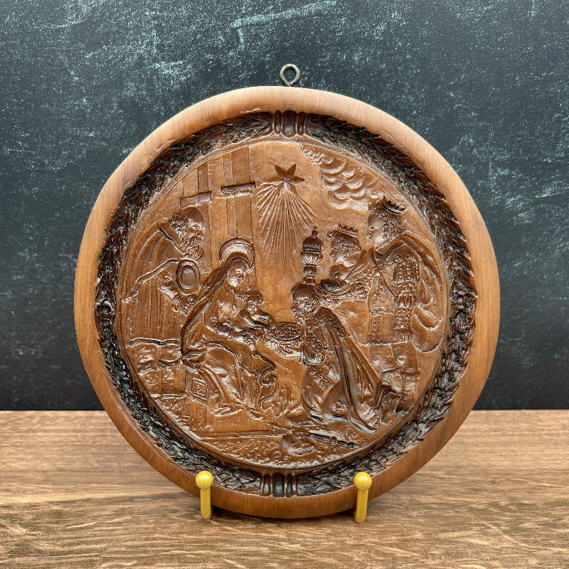 A round, hand-carved cookie mold depicting a detailed nativity scene, shown  on a split dark gray and light brown wood background.