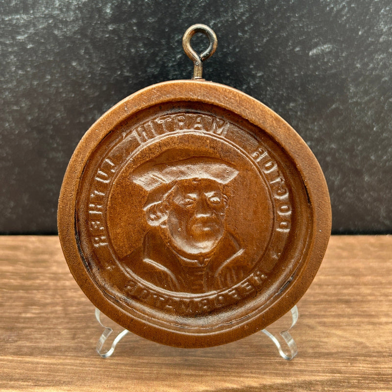 A round brown cookie cutter depicting Dr Martin Luther and raised text, displayed on a small clear stand against a dark textured background and wood surface.