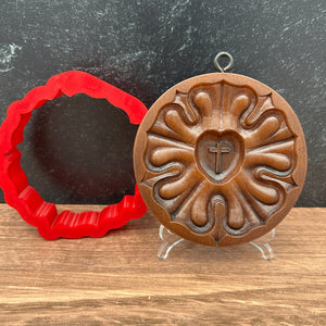 A round Luther Rose decorative mold with an embossed floral pattern and a small cross in the center, shown above a red scalloped silicone cutter on a split dark gray and wood background.