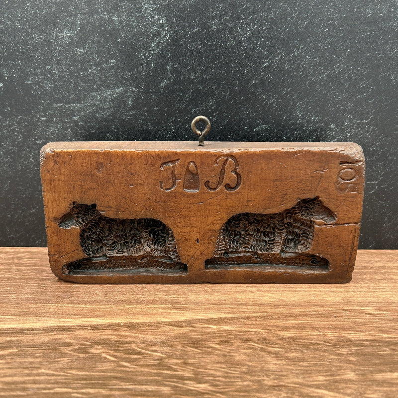 A vintage wooden cookie or soap mold with two dog cutouts and the initials 'JB' in the center, resting on a wooden surface against a dark textured background.