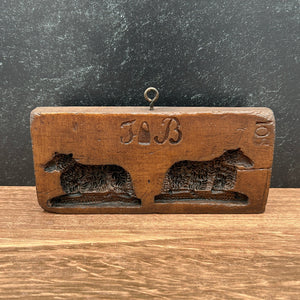 A vintage wooden cookie or soap mold with two dog cutouts and the initials 'JB' in the center, resting on a wooden surface against a dark textured background.