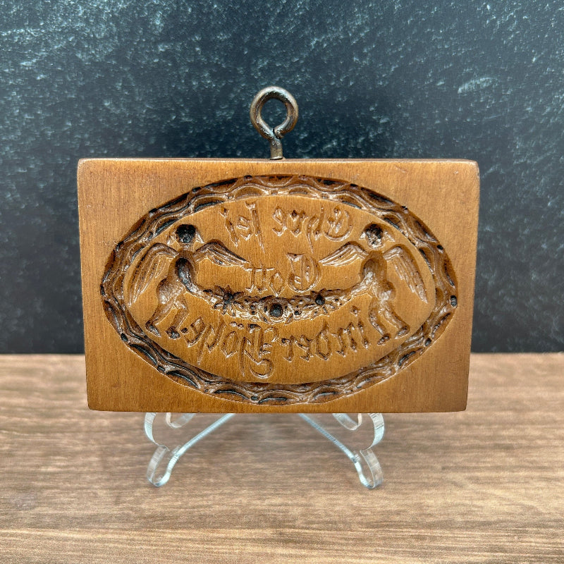 A brown cookie mold stamp with an oval embossed design featuring angels and the words "To God be the Glory" in German.  displayed on a clear stand against a split dark gray and wood-grain background.