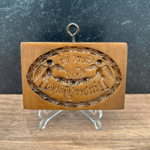 A brown cookie mold stamp with an oval embossed design featuring angels and the words "To God be the Glory" in German.  displayed on a clear stand against a split dark gray and wood-grain background.