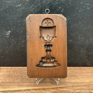 A small carved cookie mold with a hanging loop, featuring a stylized chalice and wafer details, photographed on a dark textured surface next to a wood-grain surface.