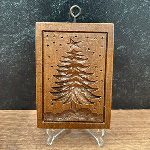 A rectangular wooden cookie mold with an embossed pine tree design, photographed from above on a dark textured surface next to a wood-grain surface, with clear acrylic stands visible on both sides.